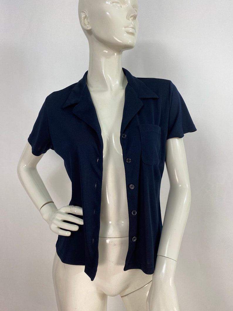 May include: A navy blue short-sleeved shirt with a collared neckline and a single chest pocket. The shirt has a button-up front and is displayed on a mannequin. The fabric appears to be a lightweight knit.