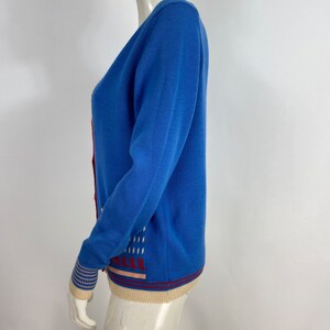 May include: A blue cardigan with a white and red geometric pattern on the front and a cream and red striped trim on the bottom and cuffs.