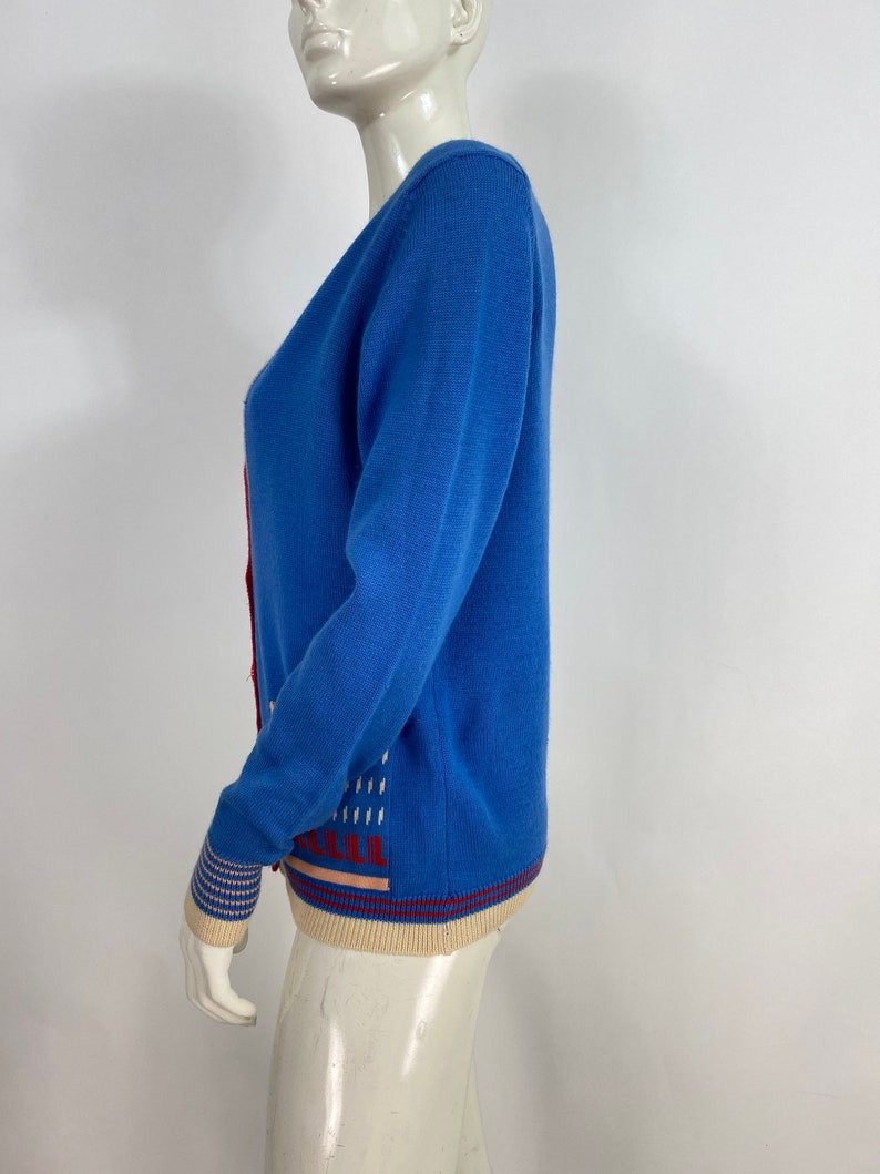 May include: A blue cardigan with a white and red geometric pattern on the front and a cream and red striped trim on the bottom and cuffs.