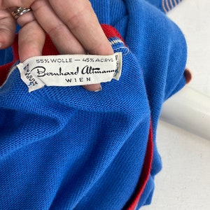 May include: A blue sweater with red and white stripes on the sleeves and a red trim around the collar. The sweater has a label that reads "55% Wolle - 45% Acryl Bernhard Altman Wien".