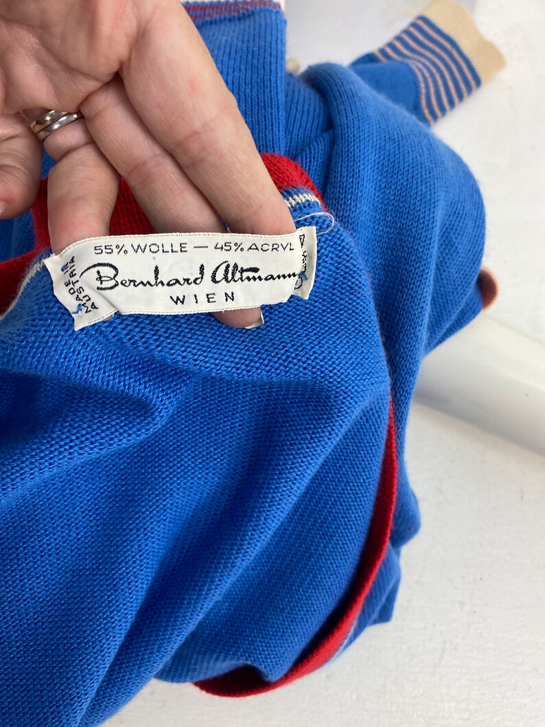 May include: A blue sweater with red and white stripes on the sleeves and a red trim around the collar. The sweater has a label that reads "55% Wolle - 45% Acryl Bernhard Altman Wien".
