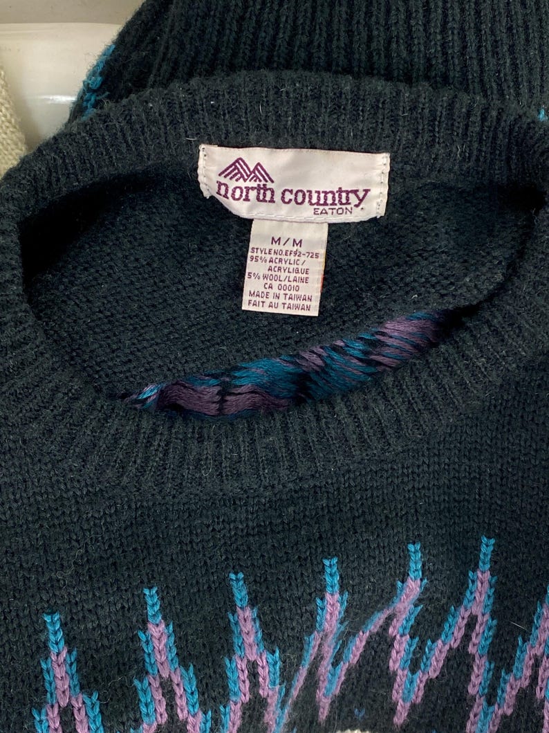 Pode incluir: Camisola preta e azul com padr&atilde;o e uma etiqueta da North Country Eaton. A etiqueta indica que a camisola &eacute; feita de 95% acr&iacute;lico e 5% l&atilde;. A etiqueta tamb&eacute;m indica que a camisola &eacute; feita em Taiwan.