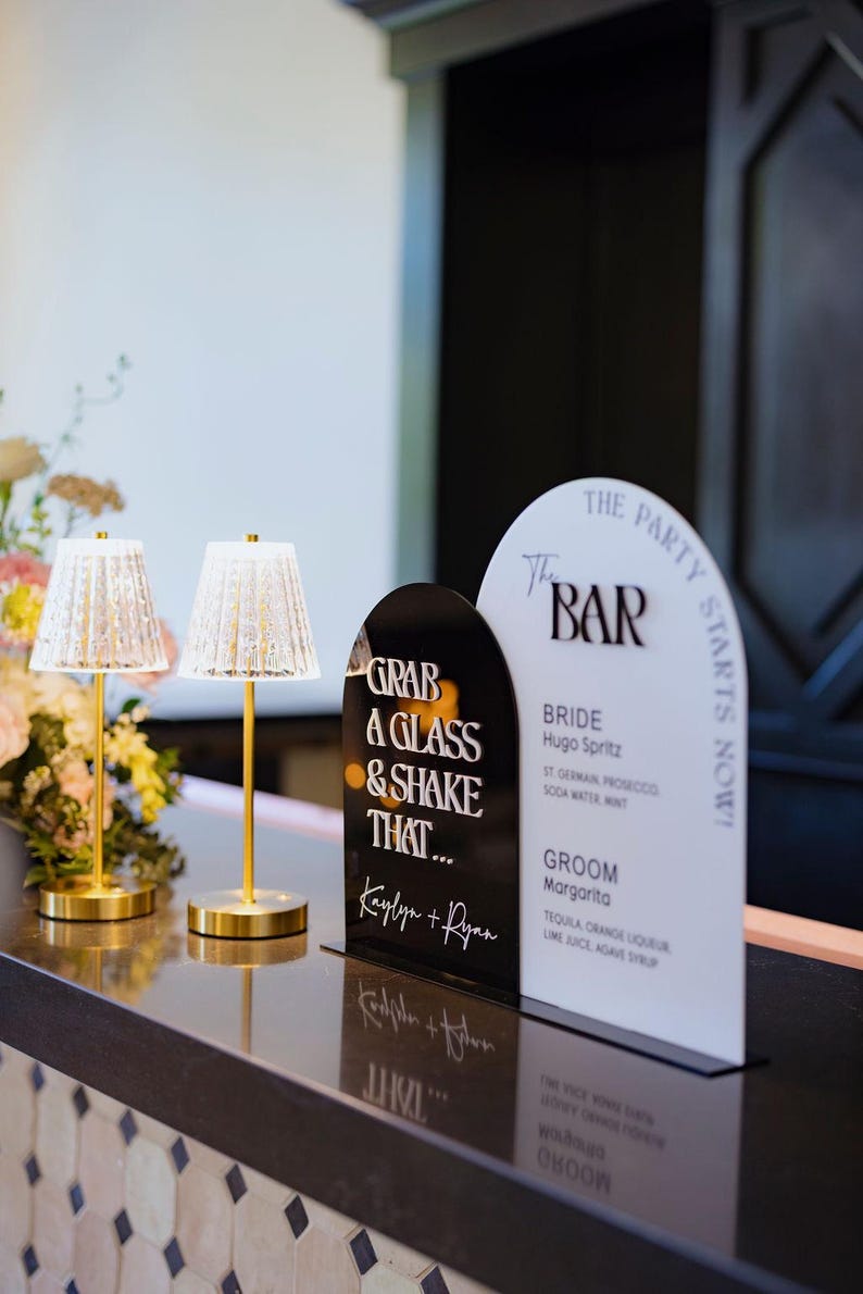 May include: Black and white sign with the text "Grab a glass & shake that... Kaylyn + Ryan" and a white sign with the text "The Bar The Party Starts Now! Bride Hugo Spritz St. Germain Prosecco Soda Water Mint Groom Margarita Tequila Orange Liqueur Lime Juice Agave Syrup".