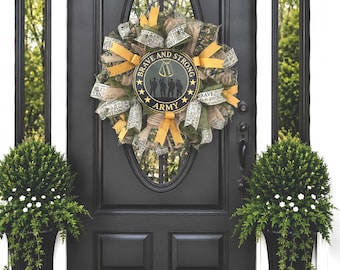 Peut inclure: Une couronne décorative composée de rubans en maille camouflage et en toile de jute avec des accents jaunes. Au centre, un panneau noir circulaire porte le texte doré BRAVE AND STRONG ARMY, avec des silhouettes de soldats et des plaques.