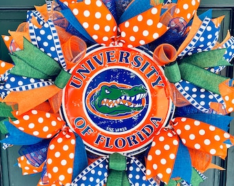 May include: A wreath decorated with orange, blue, and green ribbon with white polka dots. The wreath features a metal sign with the text "University of Florida" and an alligator logo.