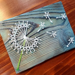 May include: A white string art dandelion with a green stem on a blue and brown wooden background. The dandelion has a circle of white string loops around the center and white string lines radiating out from the center.