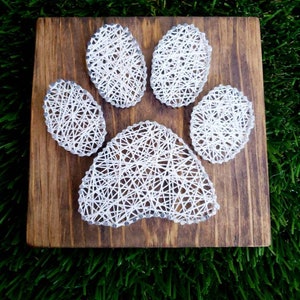 May include: A wooden plaque with a paw print design. The paw print is made of white string and is set against a dark brown wooden background. The plaque is displayed on a bed of green grass.