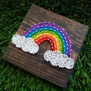 May include: A rainbow made of colorful yarn is strung on a wooden board. The rainbow has white clouds on either side.