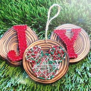 May include: A wooden Christmas ornament spelling "JOY" with red string art letters and a Mickey Mouse head. The letters and Mickey Mouse head are on round wooden slices, with a twine loop for hanging. The background is green.
