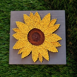 May include: A string art sunflower with yellow string on a gray wooden background.