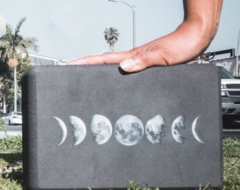 May include: A black yoga block with a moon phase design. The block is rectangular and has rounded edges. The moon phases are printed in white and are arranged in a row across the block.