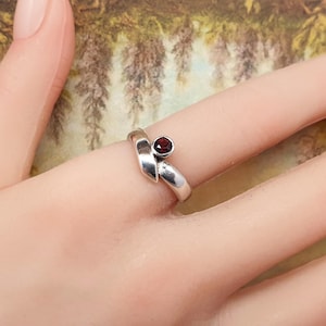 May include: A silver ring featuring a bypass design with a single round, deep red garnet gemstone set in a bezel. The band has a smooth, polished finish that curves around the finger, highlighting the contrast between the metal and the stone.