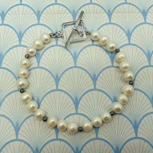 May include: A bracelet composed of creamy white pearls and small, dark grey beads. It is fastened with a silver-toned toggle clasp. The pearls are round and have a gentle sheen, giving a classic and elegant appearance.