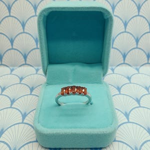 Puede incluir: Un anillo de plata con cinco piedras preciosas ovaladas de color naranja engastadas en fila. El anillo se muestra en una caja de anillo de terciopelo azul claro.