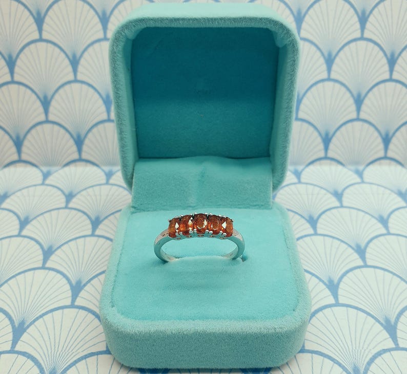 Puede incluir: Un anillo de plata con cinco piedras preciosas ovaladas de color naranja engastadas en fila. El anillo se muestra en una caja de anillo de terciopelo azul claro.