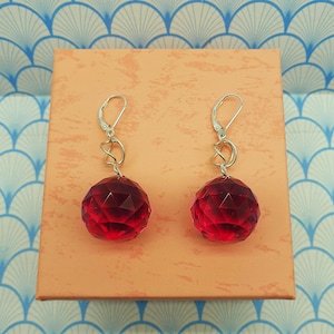 Peut inclure: Une paire de boucles d'oreilles boule en cristal rouge facetté avec des détails en métal argenté. Les boucles d'oreilles sont présentées sur une boîte pêche sur un fond bleu et blanc à motifs. Fermeture à levier.