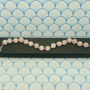 May include: A rose quartz bracelet featuring pale pink beads strung on a pink cord. The bracelet has a toggle clasp and is presented on a black box. The background has a repeating pattern.