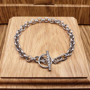 May include: A silver-toned chain link bracelet featuring a toggle clasp closure. The toggle bar is cylindrical and accented with a row of small, clear, sparkling stones. The bracelet is constructed from a series of uniform, circular metal links.