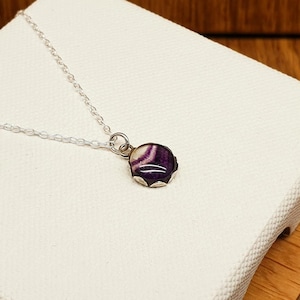 May include: A silver chain necklace featuring a small, round pendant with a polished surface. The pendant displays a distinctive pattern of deep purple and white bands, set within a simple silver-toned metal frame that secures the stone.