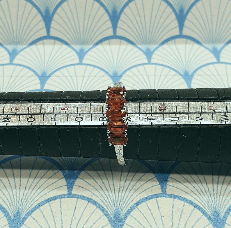 Puede incluir: Un anillo de plata con una fila de piedras preciosas de color naranja ovaladas. El anillo se mide en una regla, mostrando el tama&ntilde;o.