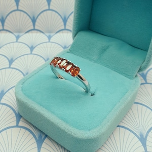 Puede incluir: Un anillo de plata con una fila de piedras ovaladas de color naranja. El anillo se muestra en una caja de anillo de terciopelo azul claro.
