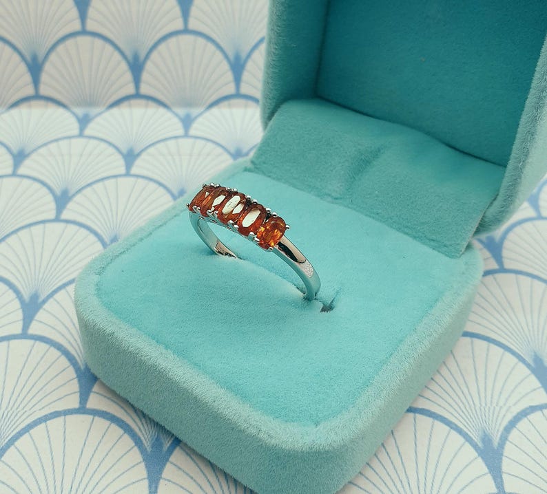 Puede incluir: Un anillo de plata con una fila de piedras ovaladas de color naranja. El anillo se muestra en una caja de anillo de terciopelo azul claro.