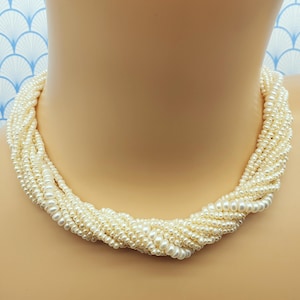 Op de afbeelding: Een crème-kleurige, meerstrengs parelketting. De ketting bestaat uit talrijke strengen kleine parels, geweven in een gedraaid ontwerp. De parels zijn dicht op elkaar gepakt, wat een volle en getextureerde uitstraling creëert. Een klassiek sieraad.