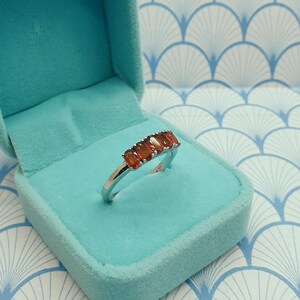Puede incluir: Un anillo de plata con una fila de piedras rectangulares de color naranja. El anillo est&aacute; en una caja de anillo de terciopelo azul claro.