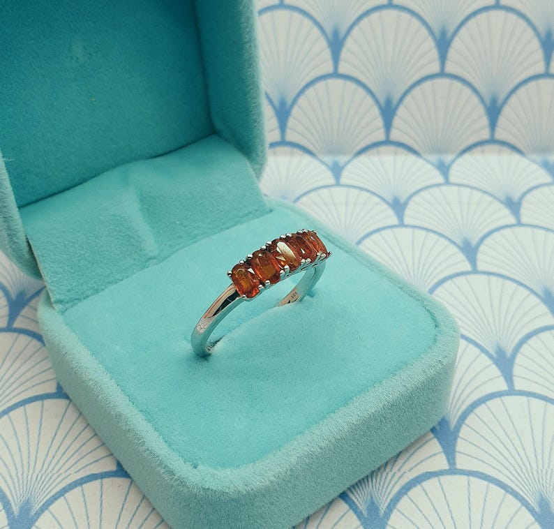 Puede incluir: Un anillo de plata con una fila de piedras rectangulares de color naranja. El anillo est&aacute; en una caja de anillo de terciopelo azul claro.