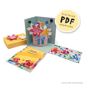 Puede incluir: Un arreglo floral de papel hecho a mano en una maceta a rayas con un lazo azul, dentro de una tarjeta desplegable. La tarjeta está abierta, revelando paneles con motivos florales y una pequeña caja de regalo con una flor de papel encima. El texto "DIGITALER PDF DOWNLOAD" es visible.