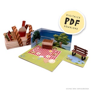May include: A papercraft picnic scene with a red and white checkered blanket, a picnic basket, and a park bench. The scene includes a bottle of wine, sandwiches, and a small box. The image has the text "DIGITALER PDF DOWNLOAD".