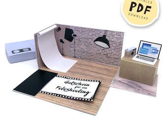 May include: A paper diorama of a photography studio, complete with a backdrop, lights, and a brick wall. A laptop and camera sit on a cardboard box. The diorama includes a gift certificate that reads "Gutschein für ein Fotoshooting."