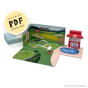 May include: A miniature paper golf course diorama with a green golf course, a small pond, and a red-roofed concession stand. The diorama includes the text "DIGITALER PDF DOWNLOAD" on a yellow circle.