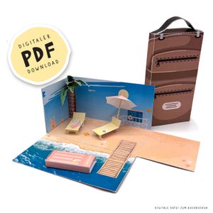May include: A papercraft diorama of a beach scene with a suitcase. The scene includes a beach, ocean, lounge chairs, and a lifeguard stand. The suitcase is brown with a handle. Text on the image says "DIGITALER PDF DOWNLOAD".