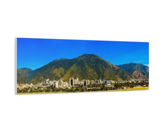 May include: Panoramic canvas print of a city skyline nestled at the base of lush green mountains under a clear blue sky. The cityscape features numerous buildings and structures, with a vibrant, natural landscape.