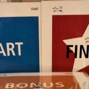 May include: Two rectangular signs with the words "START" and "FINISH". The "START" sign has a blue background, and the "FINISH" sign has a red background with a white star. Both signs have a logo in the upper left corner.