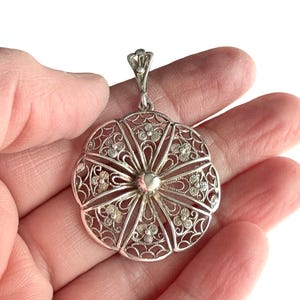 May include: A silver filigree pendant with a floral design. The pendant is round with an openwork pattern of delicate flowers and swirls. It has a small loop at the top for a chain. The pendant is held in a hand.
