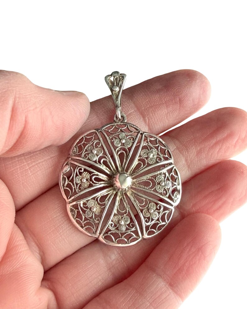 May include: A silver filigree pendant with a floral design. The pendant is round with an openwork pattern of delicate flowers and swirls. It has a small loop at the top for a chain. The pendant is held in a hand.