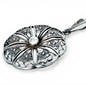 May include: A silver filigree pendant with an oval shape and a floral design. The pendant has a central, raised, circular element and a loop for a chain. The intricate openwork design is visible throughout the pendant.