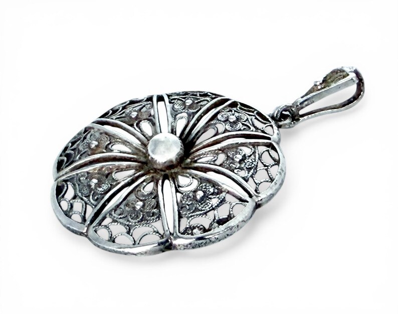 May include: A silver filigree pendant with an oval shape and a floral design. The pendant has a central, raised, circular element and a loop for a chain. The intricate openwork design is visible throughout the pendant.