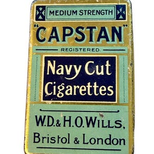Op de afbeelding: Een rechthoekig vintage metalen blik met een kleurenschema van goud, lichtgroen en donkerblauw. De tekst luidt "MEDIUM STRENGTH", "CAPSTAN", "REGISTERED", "Navy Cut Cigarettes" en "W.D. & H.O. WILLS, Bristol & London" in klassieke typografie.