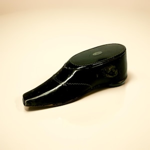 May include: A black, glossy shoe-shaped object with a pointed toe and a small heel. The shoe has decorative stitching and a textured sole. The object appears to be a decorative item or a small container.