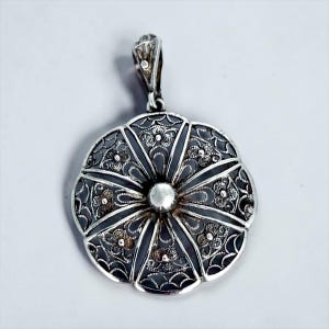 May include: A silver pendant with an intricate filigree design. The pendant is circular with a central silver bead and radiating segments. The segments are detailed with floral patterns and a decorative bail.