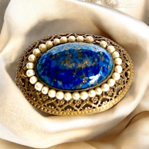 Può includere: Un broche vintage con una grande pietra ovale blu a cabochon circondata da un anello di perle finte. Il broche è incastonato in una cornice in filigrana dorata.