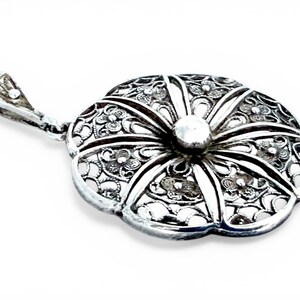 May include: A silver pendant with a floral design. The pendant is shaped like a flower with intricate filigree detailing and a small, round bead in the center. The bail is also silver and has a decorative design.
