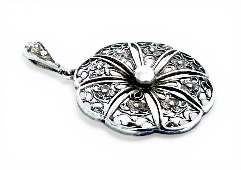 May include: A silver pendant with a floral design. The pendant is shaped like a flower with intricate filigree detailing and a small, round bead in the center. The bail is also silver and has a decorative design.
