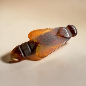 Könnte beinhalten: Ein durchscheinender, bernsteinfarbener Anhänger in Insektenform mit einem segmentierten Körper und zwei großen, flachen Flügeln. Das Material zeigt dunkelbraune und goldene Farbtöne mit einer glatten, polierten Oberfläche. Am Ende befindet sich eine kleine Metallöse.