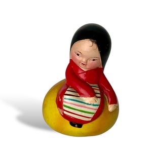 Peut inclure: Figurine en céramique peinte à la main représentant une personne assise, vêtue de noir et de rouge. La figurine est assise sur une base jaune avec un tablier rayé. C'est une pièce d'art populaire.