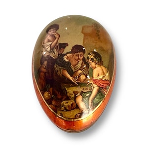 Op de afbeelding: Ovale decoratieve container met een geschilderde scène van figuren in klassieke kleding. De container heeft een rode en oranje rand en het kunstwerk toont mensen die een spel spelen. De achtergrond is een gedempt groen en bruin.