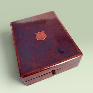 May include: A rectangular, wooden box with a glossy, dark reddish-brown finish. A small, shield-shaped emblem is centered on the lid. The box has a simple, classic design, suggesting it may be used for storage or display.